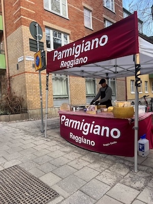 A photo of a farmer's market style tent on a city block with the words PARMIGIANO REGGIANO on the tent. At the table is what looks like a fairly handsome man in his 20s or 30s whom you could very easily believe finds easy customers by tempting passing women with free cheese to taste.