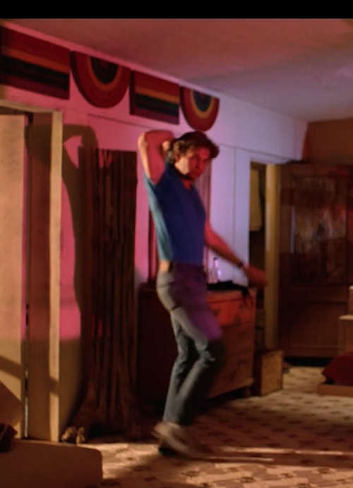 Flynn entering his own apartment. Above the doorway are several rainbow flags, some rectangular and some curved.