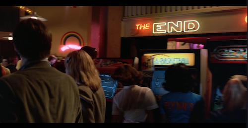 screenshot from inside Flynn's Arcade, as Lora and Alan approach where Flynn is playing Space Paranoids, they pass a sign for a game called The End